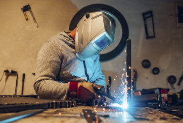 Young male welder wearing construction gloves and welding mask hardworking and welding with welding machine metal in workshop. Blue sparks. Worker in furniture factory grinding metal. Copy space.