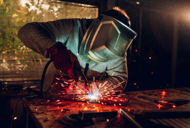 Young male welder wearing construction gloves and welding mask hardworking and welding with welding machine metal in workshop. Worker in furniture factory grinding metal and red sparks everywhere.